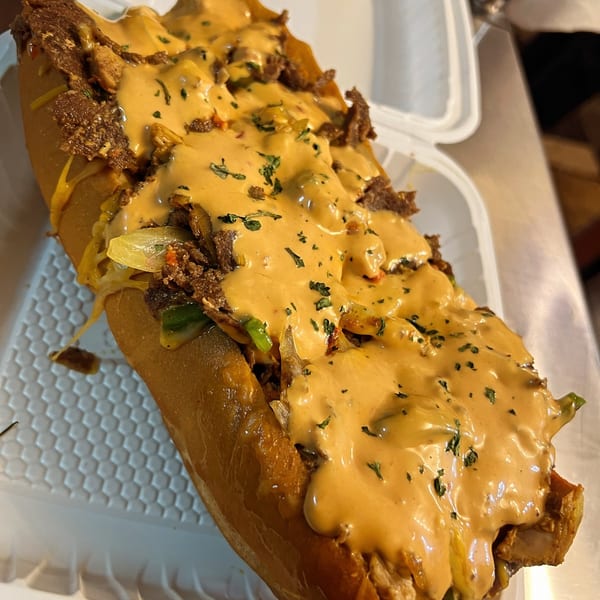 A large, cheesy American steak and cheese sub in a white takeout container.