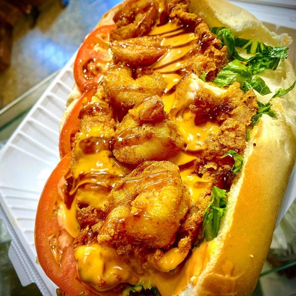 A close-up of a fried shrimp po'boy sandwich with lettuce, tomatoes, and drizzled sauces.