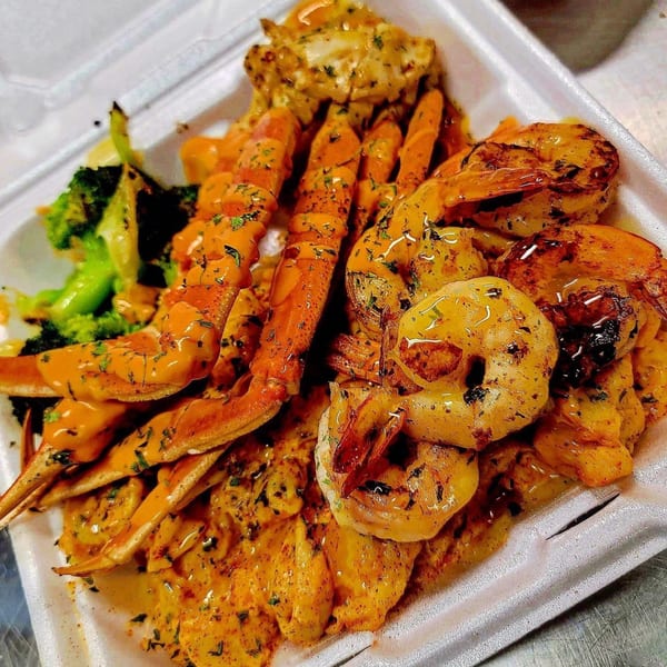 A takeout container filled with crab legs, shrimp, and broccoli, all covered in a rich sauce.